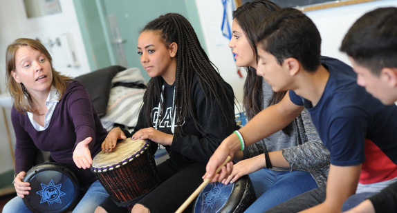 Jugendliche Schülerinnen und Schüler im Klassenzimmer mit ihrer Lehrerin am Trommeln.